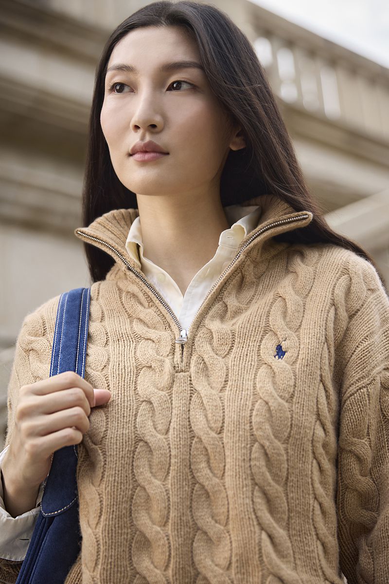 Asian woman wearing a camel cable knit quarter zip sweater and holding a blue bag outdoors