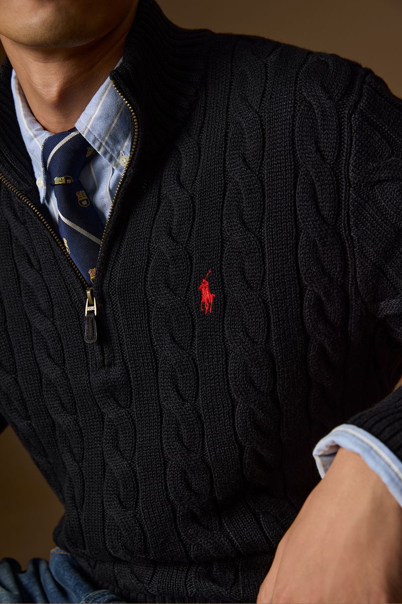Man wearing a black cable-knit quarter-zip sweater with an embroidered red logo over a light blue collared shirt and patterned tie.