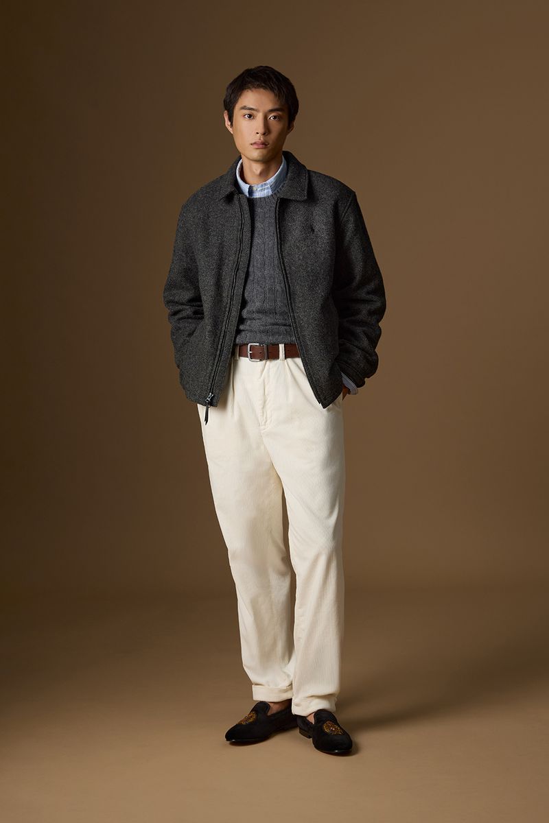 Man standing in a charcoal zip jacket, grey knitwear, cream pleated trousers, and black embroidered loafers against a tan backdrop.