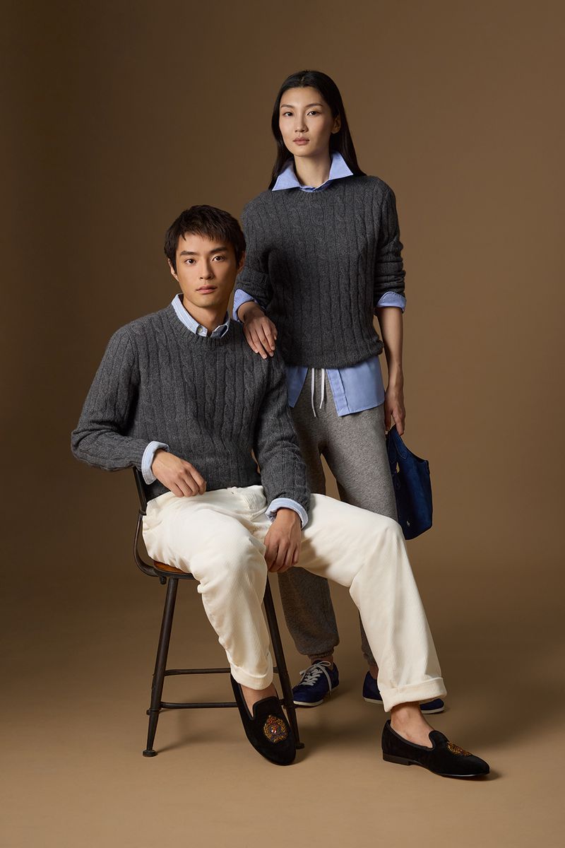 A man and a woman in matching grey cable-knit sweaters and light blue shirts pose against a warm tan backdrop.