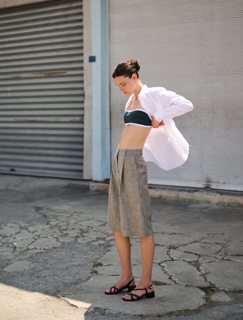Young woman in white button-down shirt, green top, and culottes stands in an urban setting with a corrugated metal background.