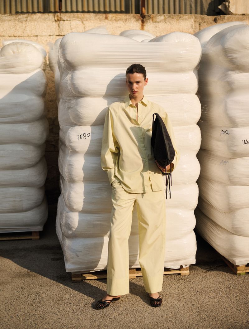 Fashion shot of a woman in a light yellow pantsuit holding a black bag, standing in front of a stack of white wrapped bales outdoors.