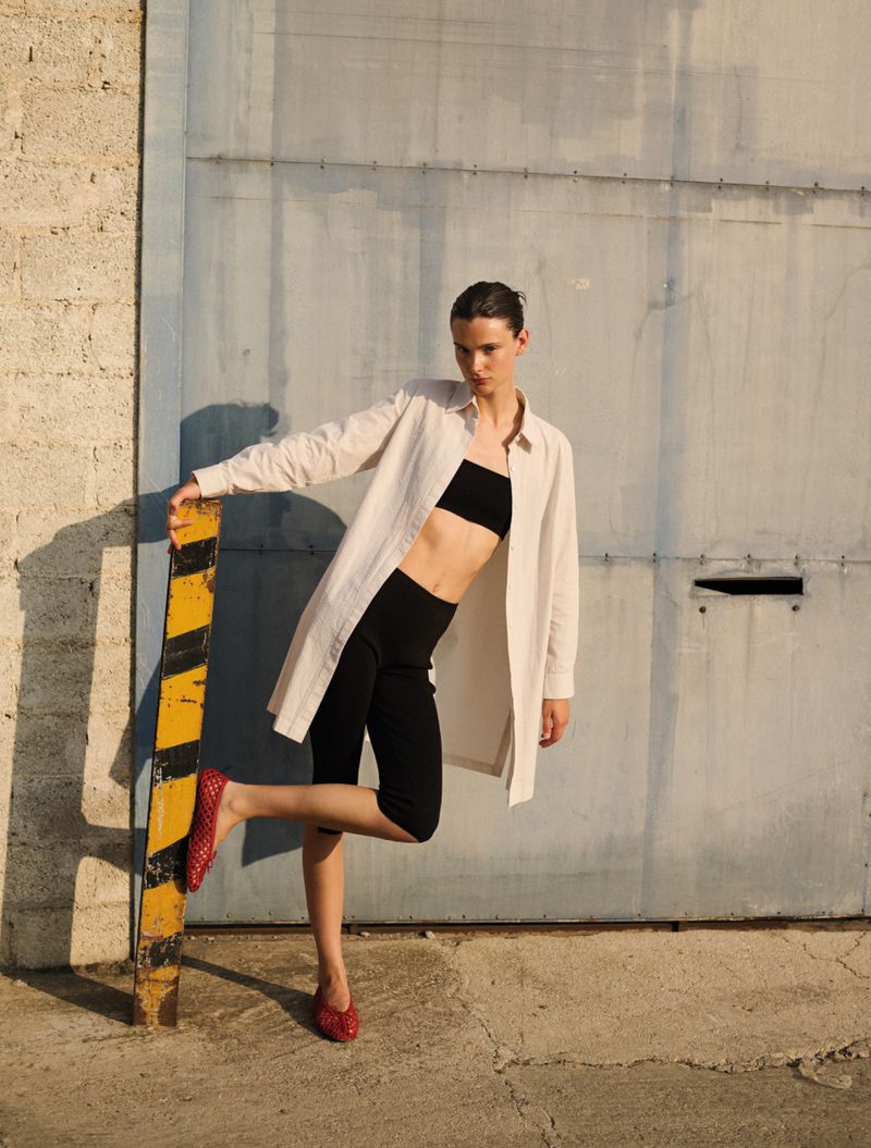 A young female fashion model poses outdoors in an unbuttoned linen shirt, black crop top, dark capri pants, and red woven flats.