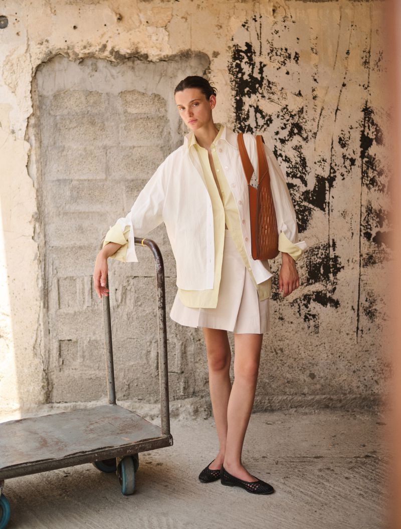 A fair-skinned adult woman with a tan leather bag poses in a neutral skirt and white shirt, leaning on a metal luggage trolley.