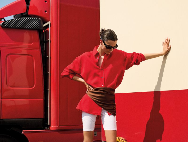 Fashionable woman poses in red blouse, white shorts, and sunglasses next to a large red truck and cream-colored wall outdoors in daylight.