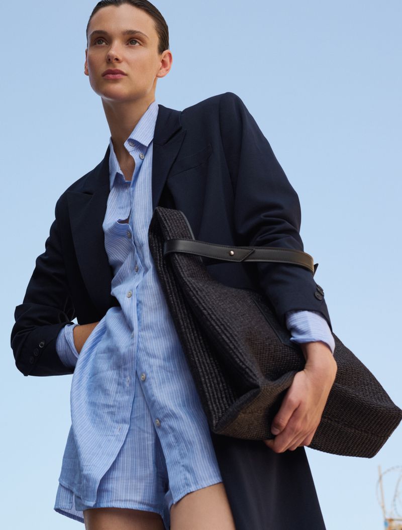 A woman poses in a blue shirt and blazer, holding a woven tote bag against a bright blue sky backdrop. The shot is low angle.