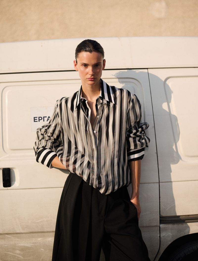 Fashion portrait of a model in a striped blouse and dark slacks, posed against a white van in soft, neutral daylight.