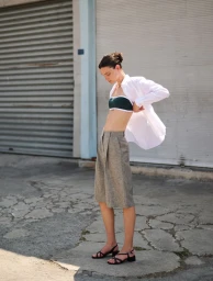 Sara Dijkink posing in an open white shirt, dark bandeau top, tailored grey bermuda shorts, and black strappy sandals outdoors.