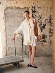 Model Sara Dijkink wearing layered white and yellow shirts with a brown handbag standing beside a metal cart against a weathered wall