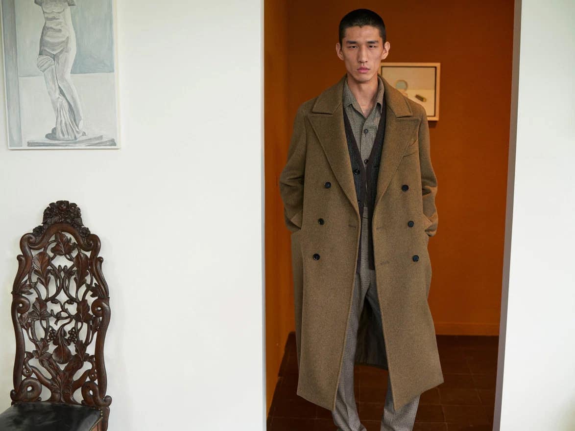 Man wearing a brown double-breasted coat standing in an interior space with white walls and artwork.