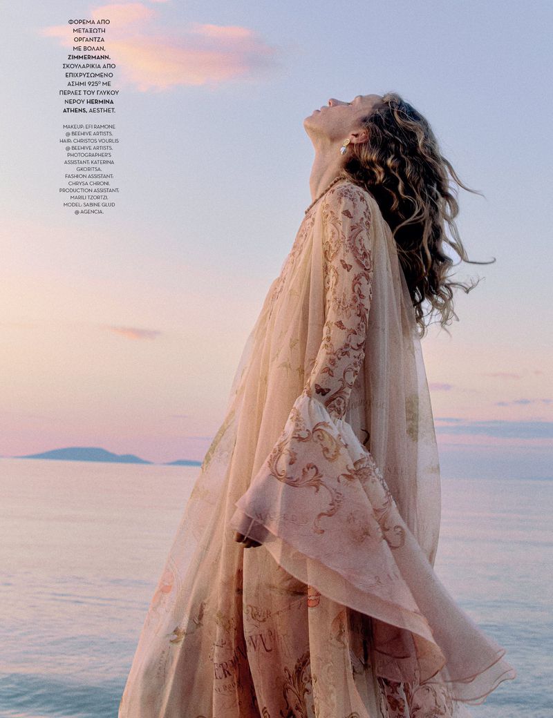 Woman in flowing patterned dress gazes up towards the sky with a coastal view and pink and blue sky in the background.