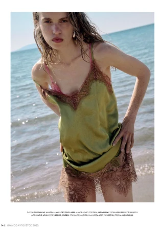 A woman in a green lace-trimmed dress stands on a beach with a neutral expression, featuring a blend of fashion and natural beauty.
