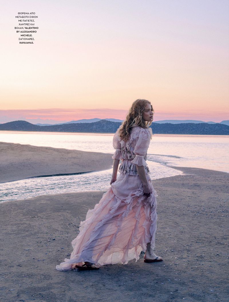 Woman in a long flowing dress walking on a sandy beach at sunset with mountains in the background under a pastel sky. Fashion shoot.