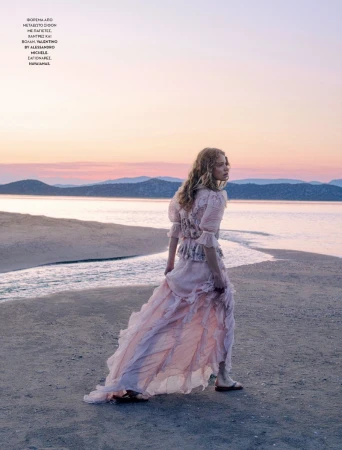 Woman in a long flowing dress walking on a sandy beach at sunset with mountains in the background under a pastel sky. Fashion shoot.