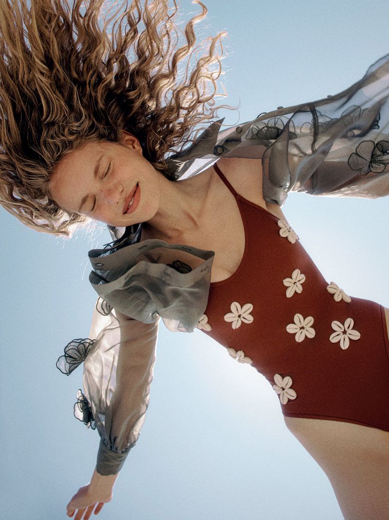 Woman in a rust-colored floral swimsuit lies against a blue sky, wearing a sheer floral top, photographed from a low angle on a sunny day.