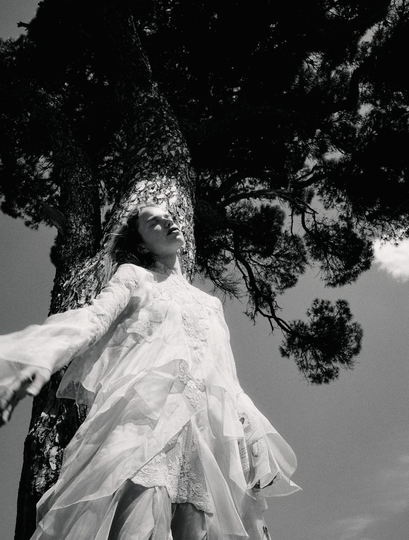 Low-angle black and white shot of model Sabine Glud in a layered white lace dress leaning against a large tree trunk.