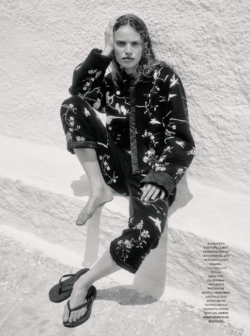 Monochrome portrait of a woman in a patterned jacket and pants sitting on a white surface with sandals beside her.