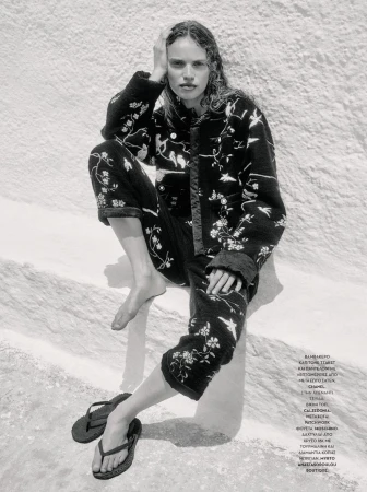 Monochrome portrait of a woman in a patterned jacket and pants sitting on a white surface with sandals beside her.