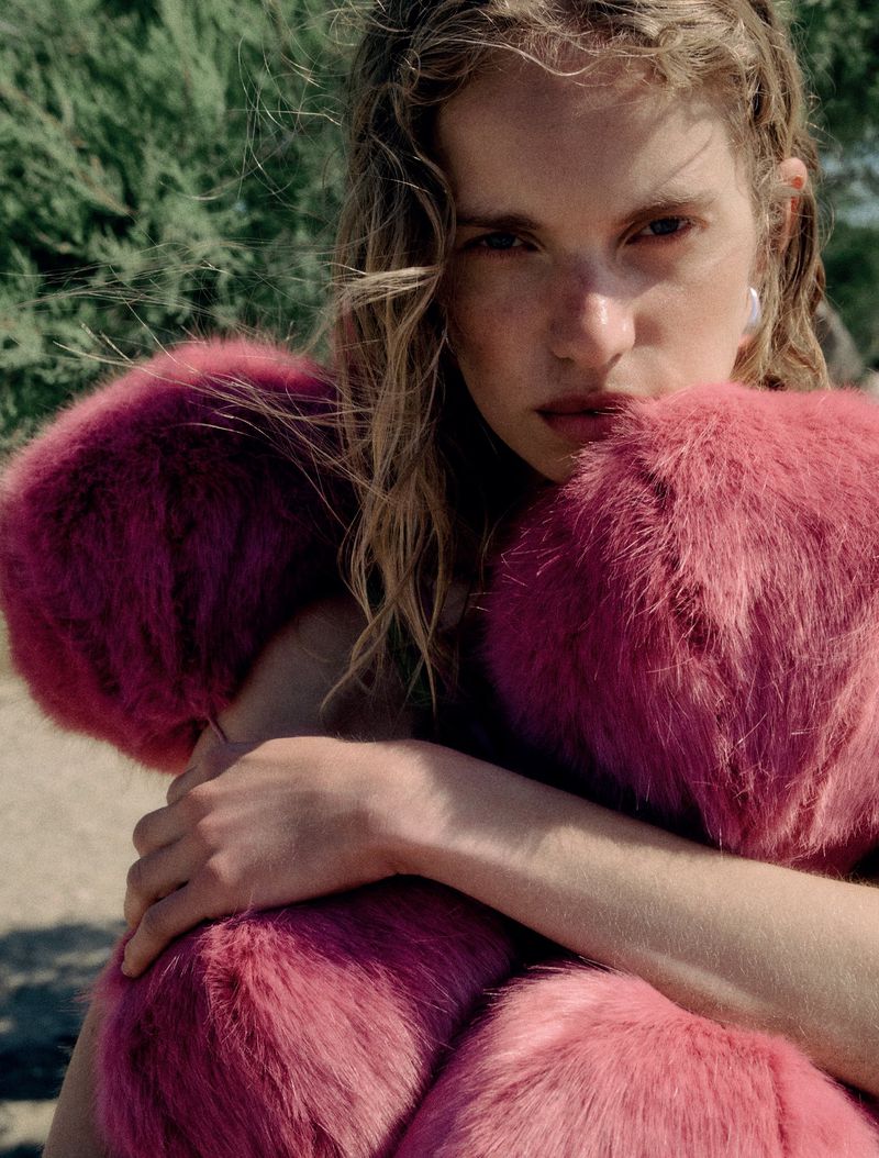 Woman in pink faux fur coat poses outdoors in front of green foliage, portrait-style, fashion editorial shot, soft lighting and natural setting.