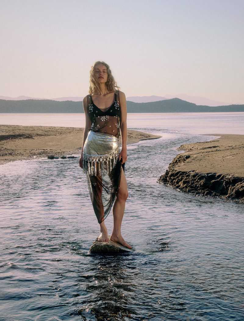 A woman in a sheer top and silver skirt stands in a stream at the beach with mountains in the distance on a sunny day.