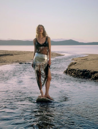 A woman in a sheer top and silver skirt stands in a stream at the beach with mountains in the distance on a sunny day.