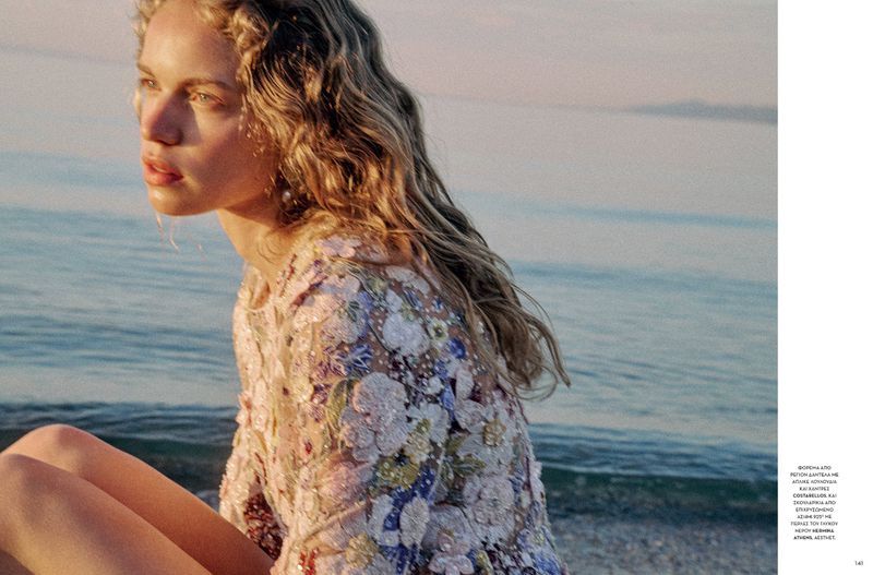 A young adult female sits in soft light near the ocean, wearing a floral-patterned dress with blonde wavy hair, in a fashion setting.