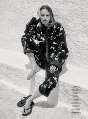 Model Sabine Glud seated against a textured white wall wearing patterned dark clothing for Vogue Greece sea flowers editorial.