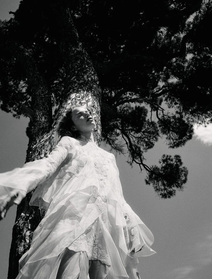 Low-angle black and white shot of model Sabine Glud in a layered white lace dress leaning against a large tree trunk.