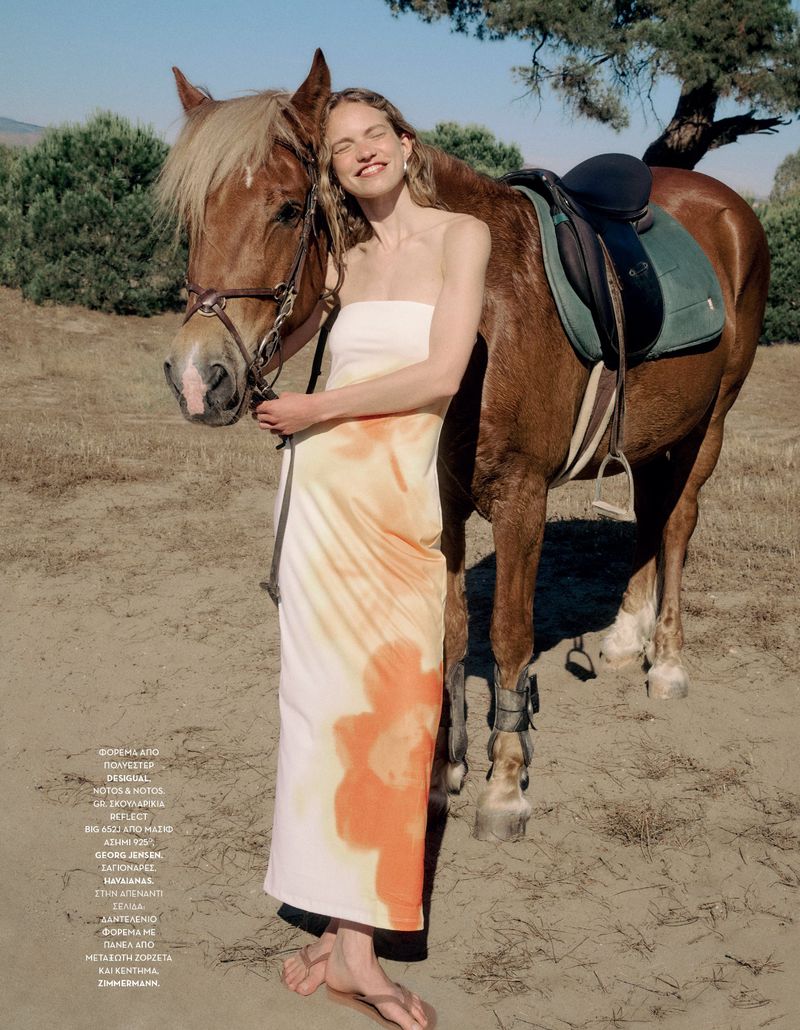 Model in a strapless floral-print dress hugging a brown horse outdoors in a dry landscape for Vogue Greece