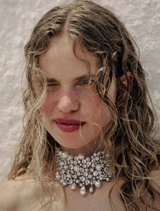 Model Sabine Glud wearing an ornate pearl and crystal choker necklace with wavy hair against a textured white wall.