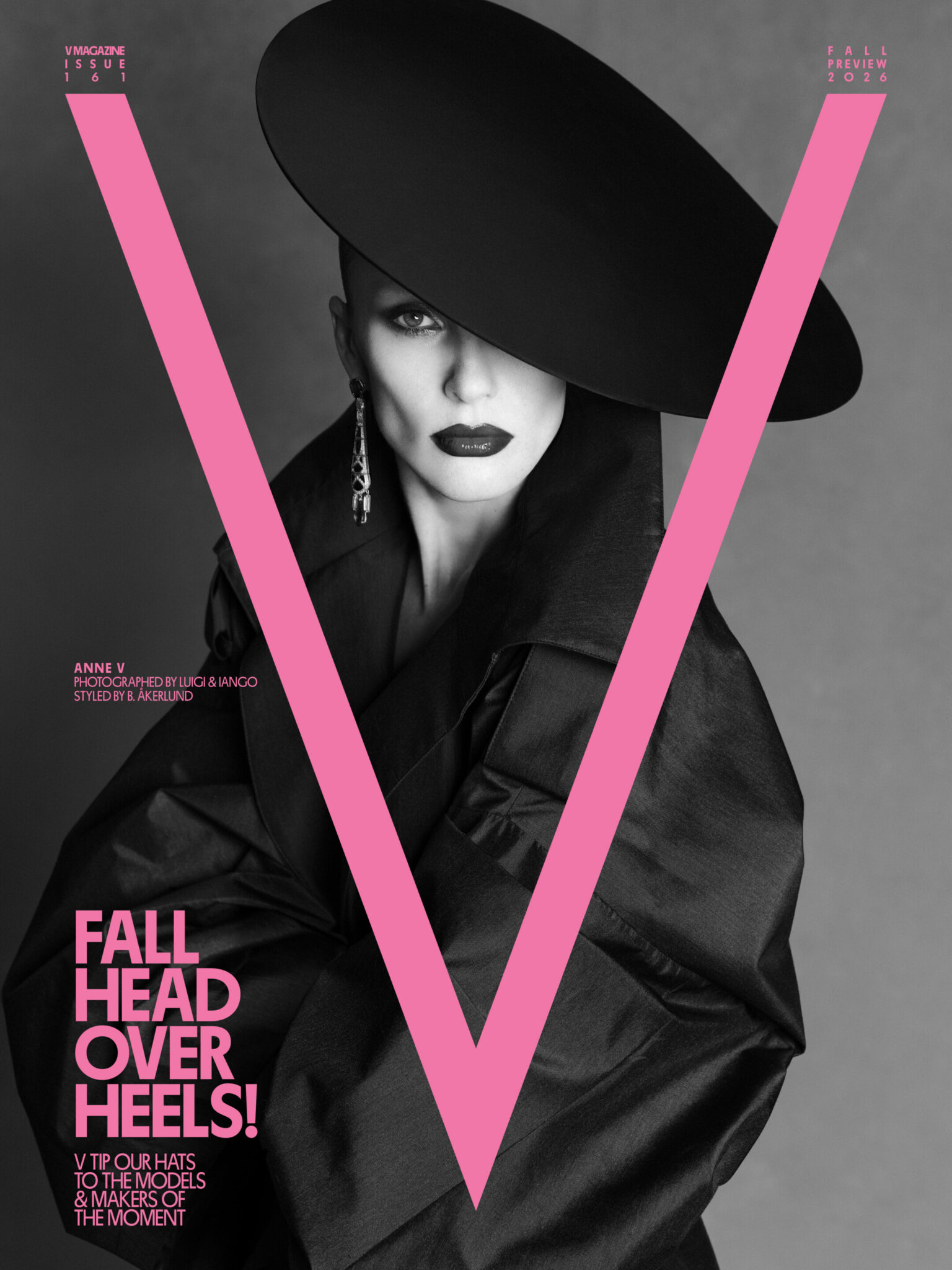 Model Anne Vyalitsyna wearing a wide-brim tilted black hat and high-collar black coat framed by a large pink V letter graphic.