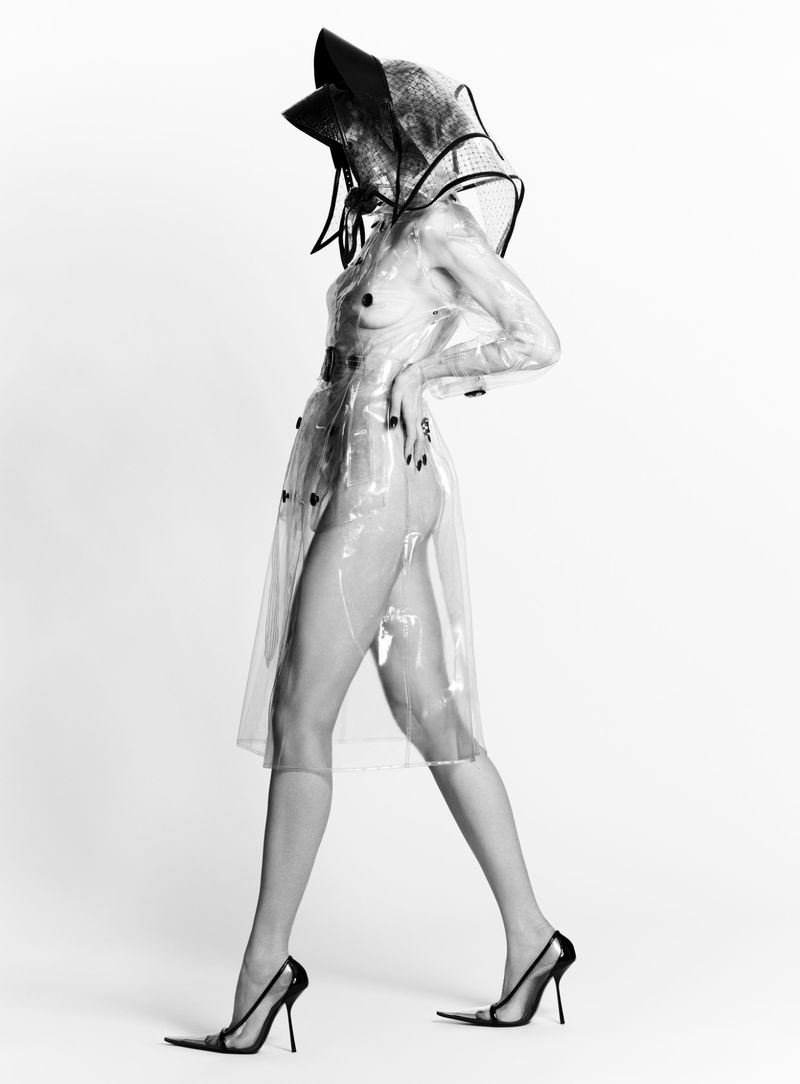 Anne Vyalitsyna wearing a sheer transparent trench coat, sculptural black hat, and stiletto heels against a white studio background.