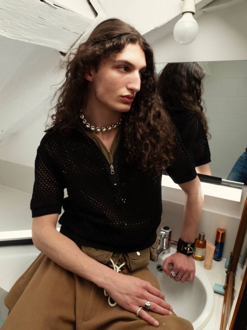 A long-haired adult male sits on a bathroom sink wearing a black top, brown skirt, and beaded necklace, with bathroom fixtures visible.