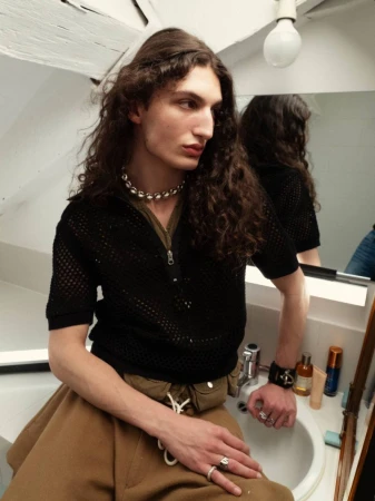A long-haired adult male sits on a bathroom sink wearing a black top, brown skirt, and beaded necklace, with bathroom fixtures visible.