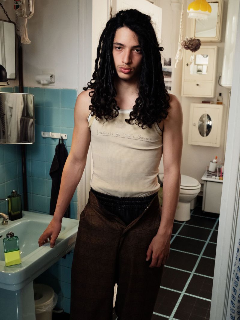 Male in a bathroom wearing a tank top and unbuttoned brown pants, standing near a light blue sink, with bathroom cabinets and accessories visible.
