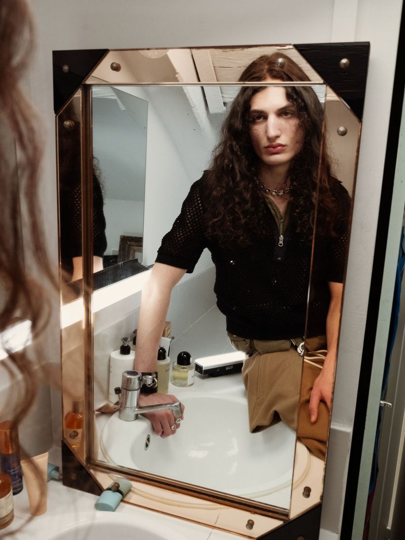 Reflection in a bathroom mirror of a person with long brown hair wearing a black shirt and tan pants, standing near a white sink.