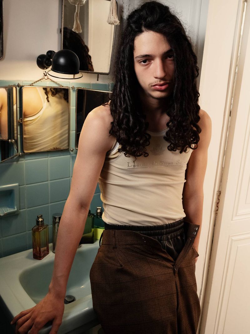 Young adult in a tan tank top and unbuttoned brown pants poses in a bathroom with a vintage sink, mirror, and toiletries.