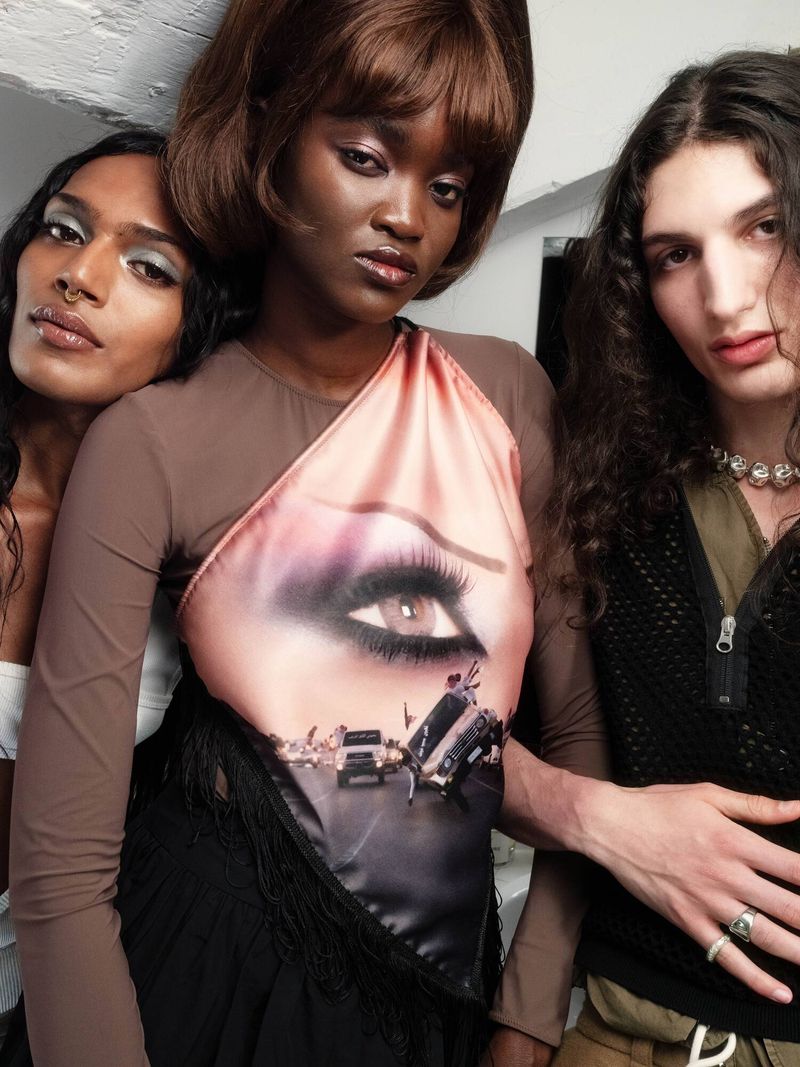 Three diverse individuals pose indoors. One with an eye-graphic top, another with a white top, and one with curly hair in a casual setting.