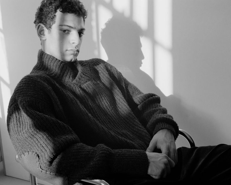 Monochrome portrait of a young man seated in a chair wearing a dark ribbed turtleneck sweater with window shadows on the wall behind him.