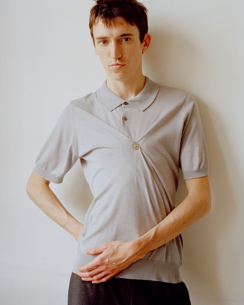Young man posing in a light grey short-sleeved gathered knit polo shirt against an off-white studio background.