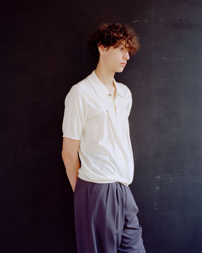 A young man with curly hair wearing an off-white short-sleeved polo shirt and pleated dark trousers leans against a dark wall.