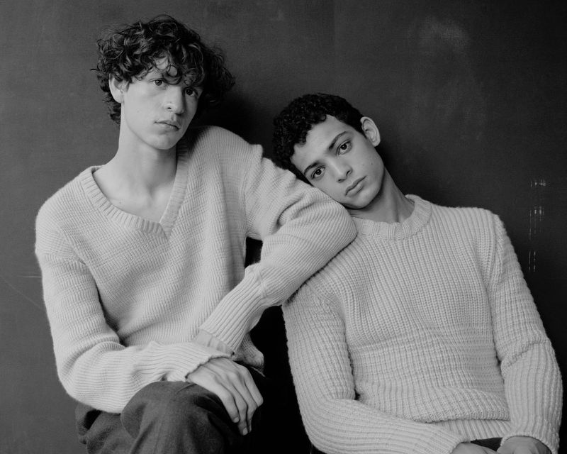 Two young male models in light ribbed knit sweaters posing closely together against a plain dark background in black and white.