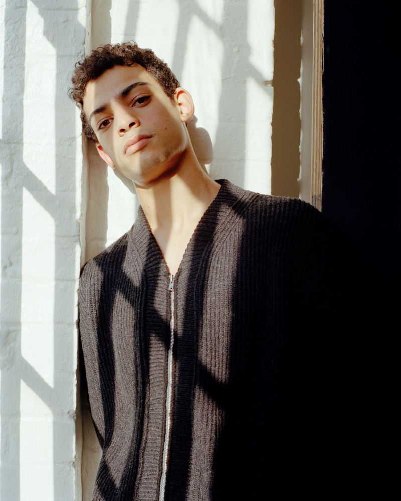 Man in a black zip cardigan leaning against a sunlit white brick wall with window shadows