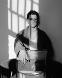 Black and white portrait of a man seated backward on a wire chair in a dark jacket and white tank top under geometric window shadows.