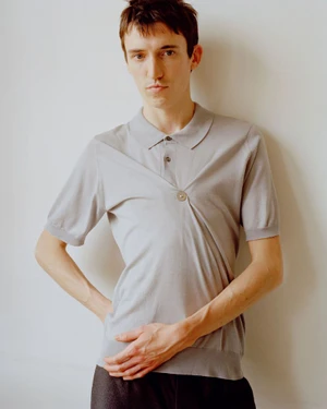 Young man posing in a light grey short-sleeved gathered knit polo shirt against an off-white studio background.