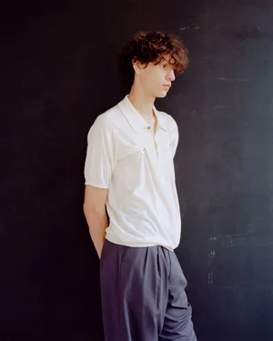 A young man with curly hair wearing an off-white short-sleeved polo shirt and pleated dark trousers leans against a dark wall.