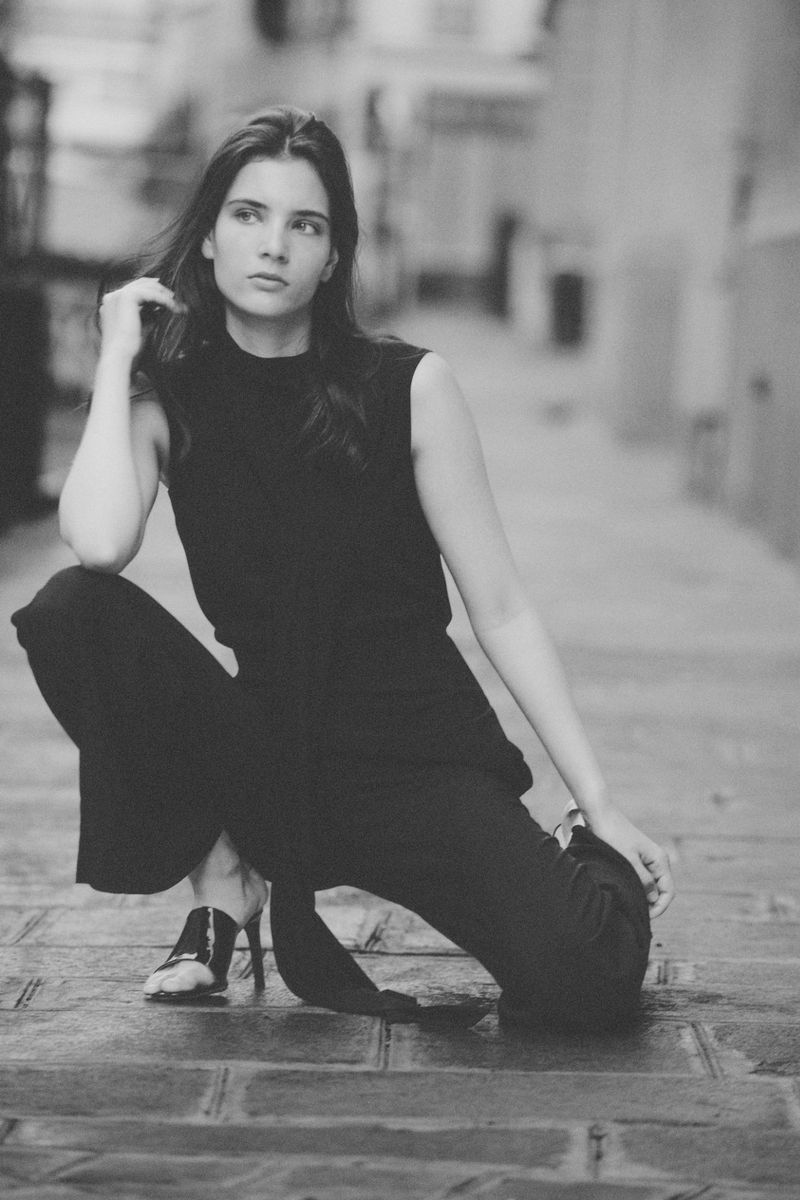 Monochrome portrait of a woman crouching on a paved street wearing a black sleeveless top, matching trousers, and heeled mules.