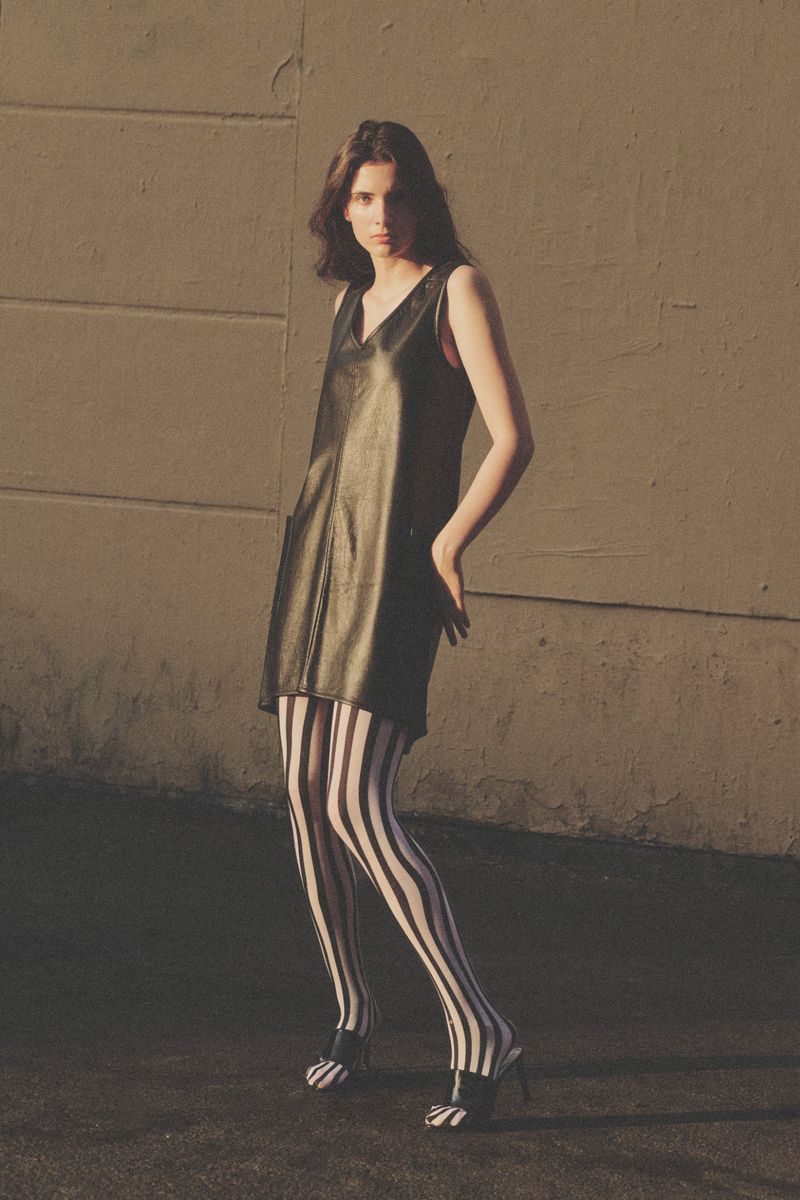Model posing outdoors in a black sleeveless mini dress and bold black-and-white vertically striped tights against a concrete wall.