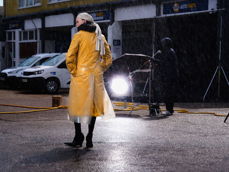 Model standing outdoors in the rain wearing a long yellow raincoat and headscarf with parked vans and a lighting rig behind them