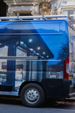 Side profile of a parked blue wrapped commercial van with roof racks on a residential street in front of a Victorian building.